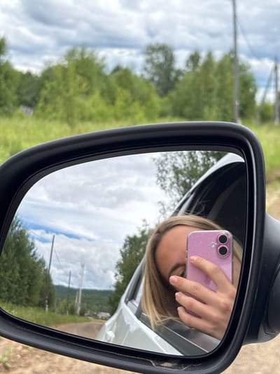A woman takes a selfie in a car's side mirror. She is leaning out of the car window, holding a pink phone. Her blonde hair is visible, and she is looking at the phone. The car appears to be on a dirt road in a rural area. The mirror reflects a cloudy sky, green trees, and a road winding through a hilly landscape. In the distance, there are some buildings and utility poles, possibly indicating the outskirts of a settlement like Sedanovo, Russia. The scene is set in a natural environment with vegetation and open sky.