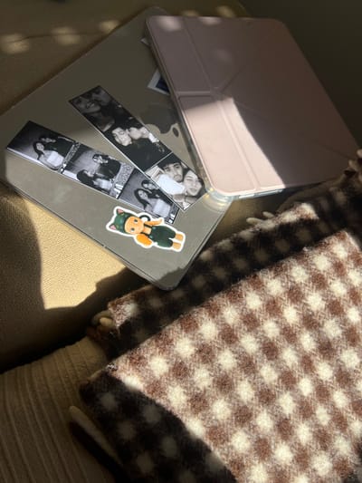 An image shows a pink-covered iPad atop a silver MacBook. Photo strips and a cartoon cat sticker adorn the laptop. A brown and white checkered blanket is beside the electronics. All items are resting on a light brown, textured surface. Sunlight illuminates portions of the composition, creating shadows.