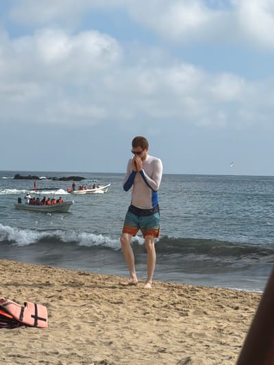 A man wearing a long-sleeved white and blue shirt and colorful shorts stands on a sandy beach near the ocean. He appears to be preparing to enter the water. A life vest lies on the sand nearby. Several boats filled with people are visible in the ocean behind him. The sky is partly cloudy.