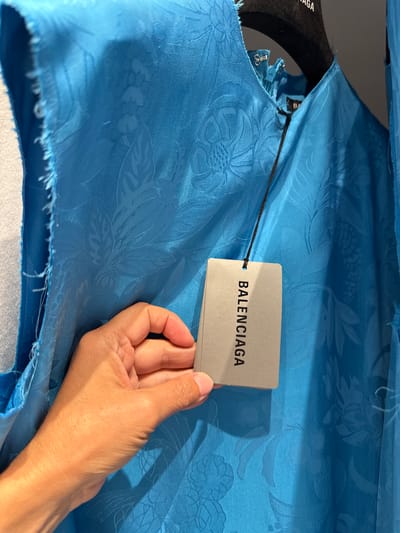 The image shows a close-up of a blue textured dress hanging on a black hanger. A hand is holding a Balenciaga tag in front of the dress. The dress has a floral pattern embossed into the fabric. The background is out of focus and appears to be a store interior. The scene is likely in a Balenciaga store, possibly in La Roca del Vallès, Spain, which is known for its luxury outlet shopping.