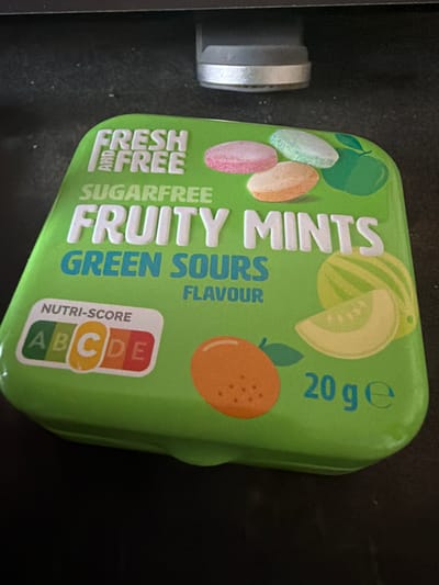The image displays a green tin of "FRESH AND FREE SUGARFREE FRUITY MINTS GREEN SOURS FLAVOUR". The tin is oriented at an angle, showing the top and right side. It features illustrations of various fruits, including pink, light blue, and orange lozenges, a green apple, a watermelon, a slice of melon, and an orange. A Nutri-score label with the letter "C" highlighted in yellow is visible on the left side of the tin, next to the letters "A", "B", "D", and "E" in their respective colors. The text "20 g e" is printed on the right side of the tin. The tin is resting on a dark, textured surface, likely a table, with a blurred background that includes a portion of a silver and grey object. The lighting suggests an indoor setting. There are no people or discernible city elements, and therefore no connection to Torremolinos, Spain, can be made from the visual information provided.