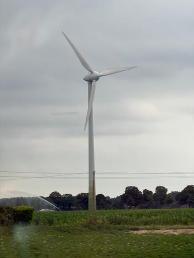 A single wind turbine stands tall in a field under a cloudy sky in Kerken, Germany. The turbine has three white blades, one of which is angled upwards, and a white cylindrical tower. In the foreground, a sprinkler system is watering a field of green crops, likely corn, with a stream of water arcing across the frame. Power lines are visible stretched across the middle ground. In the background, a line of trees and some buildings can be seen, suggesting a rural or semi-rural setting. The overall impression is one of agricultural activity and renewable energy generation.