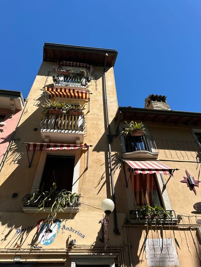 The image shows a close-up, low-angle view of the facade of a building in Desenzano del Garda, Italy, under a clear blue sky. The building is tall and narrow, with a pale yellow stucco exterior. It features multiple balconies adorned with potted plants and red and yellow striped awnings.

On the leftmost section of the building, a pink-colored neighboring structure is partially visible. The main building has several windows, some with decorative railings and others covered by awnings. On the lower part of the building, a circular emblem or crest is painted on the wall, with lettering that appears to read "Dante Andreina." Below this, a small plaque with the number "68" is visible.

To the right, a stone chimney with terracotta tiles protrudes from the roofline. Another balcony with plants and an awning is present on this side. A decorative metal star-shaped object is mounted on the wall to the right of a window. Below this, a stone plaque with Italian text is affixed to the facade. The scene suggests a residential or commercial building, possibly a guesthouse or hotel, given the balconies and decorative elements. The lighting indicates it is daytime, with strong sunlight casting shadows on the building.