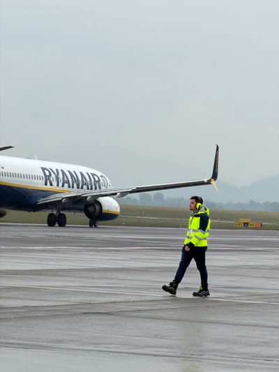 The image shows a Ryanair Boeing 737 aircraft on a runway at Orio al Serio International Airport, near Bergamo, Italy. A ground crew worker wearing a high-visibility yellow vest and headphones stands near the aircraft's tail, seemingly conducting pre-flight checks. The sky is overcast, suggesting a cloudy day. The runway markings are visible, indicating the direction of the aircraft's movement. The overall setting is a standard airport environment.