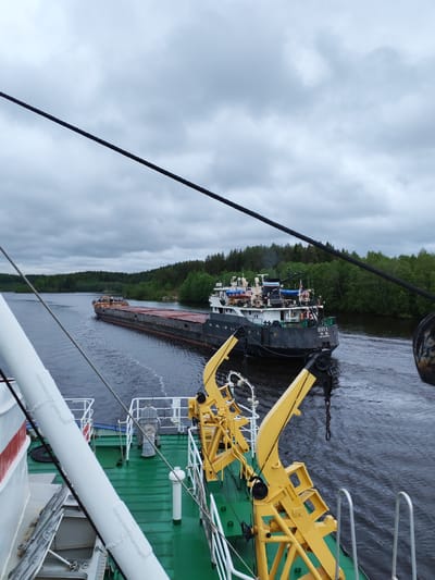 The image shows a river scene, likely in the city of Depo, Russia, judging by the Cyrillic text on the stern of the vessel in the distance. The foreground is dominated by the green deck of a boat with a yellow crane. Behind this, a long, black barge being towed by a tug boat is visible, moving along the river. The river is flanked by lush green trees on both sides. The sky is overcast with gray clouds.