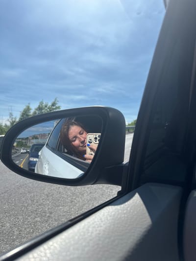 In the reflection of a car's side mirror, a young woman is visible. She is leaning out of the car window, holding a smartphone to take a selfie. Her eyes are closed, and her head is tilted upwards, suggesting a moment of enjoyment or relaxation. Her right hand, adorned with blue nail polish, is holding the phone. The phone case features a cartoonish design. The background reflected in the mirror shows a road with other vehicles, trees, and buildings. The overall scene appears to be a sunny day, with a clear sky visible. The image was taken from inside a vehicle, looking outwards, with the car's interior components, like the door frame and mirror housing, visible in the foreground. The location is identified as the city of Krutoberegovoy, Russia.