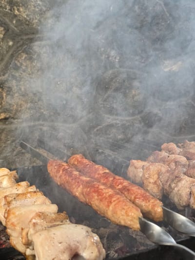 In Pidhorodne, Ukraine, a close-up shot captures the grilling of meat on skewers over a fire. The foreground features skewers laden with pieces of chicken and two elongated, ground meat kebabs. Behind them, more cubes of meat are being cooked. Wisps of smoke rise from the grill, partially obscuring the rough, grey stone background. The lighting suggests an outdoor setting, likely a backyard or a dedicated barbecue area.