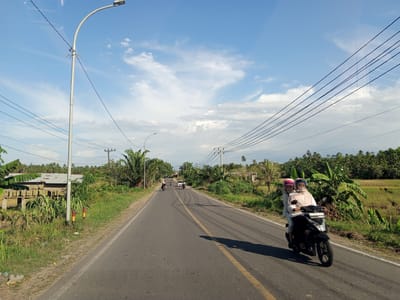 The image depicts a rural road scene in Payabeunyot, Indonesia. The road, marked with a single yellow centerline, stretches into the distance under a partly cloudy sky. Two people are riding a black motorbike on the right side of the road. The rider and passenger both wear helmets. Further down the road, a car and two more motorbikes are visible. The roadside is lined with dense vegetation, including banana trees and tall grasses. Utility poles run along the road, carrying power lines overhead. Buildings with corrugated iron roofs are visible in the background.