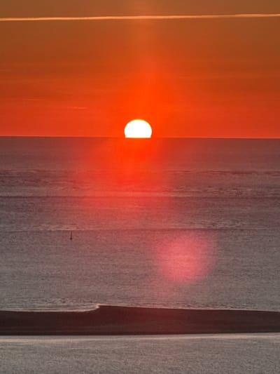 The image captures a sunset over the ocean, likely near La Teste-de-Buch, France, a coastal town known for its beaches and pine forests. The sun, a bright white orb, is partially submerged below the horizon, casting a warm orange and red glow across the sky and water. The sky is a gradient of vibrant orange and red hues, with a thin, bright white line near the top, possibly a contrail or high-altitude cloud. The ocean surface is calm, with subtle horizontal ripples reflecting the sunset's colors. A small, dark, vertical marker, possibly a buoy, is visible in the middle ground of the water. In the foreground, a dark, horizontal landmass, potentially a sandbar or a low-lying island, separates the body of water from another calmer, reflective surface below. Two large, soft, pinkish-red oval shapes are present in the lower half of the water, likely lens flares or reflections of the sunset. The overall scene evokes a serene and picturesque coastal sunset.