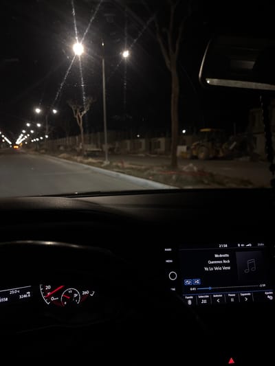 The image depicts a nighttime scene from the interior of a stationary vehicle in Komchén, Mexico. The perspective is through the front windshield, looking forward down a street illuminated by numerous streetlights.
**Vehicle Interior (Foreground):**
The lower portion of the image shows the car's dashboard and instrument panel, predominantly dark.
* On the left, a digital instrument cluster is visible, indicating a speed of 0 km/h, a fuel gauge that is approximately half full, an odometer reading of 3246.6, and an ambient temperature of 23.0°C.
* On the right, an infotainment screen displays the time as 21:58. It shows "Moderatto" as the band and "Queremos Rock" and "Ya Lo Vela Venir" as songs, with "Ya Lo Vela Venir" currently playing at the 0:45 mark of an approximately 2:59 duration. Playback controls are visible at the bottom of the screen.
* A portion of the steering wheel is visible on the left, and a rearview mirror assembly hangs from the top right, partially obscured. A red hazard light indicator is barely visible at the very bottom center.
**Exterior Scene (Midground/Background):**
The area outside the vehicle is dark, illuminated by artificial streetlights.
* A paved road, likely asphalt, stretches into the distance, with a curb defining its edge.
* Numerous streetlights with bright, white light are spaced along the road, creating starburst effects due to the camera's aperture or lens characteristics.
* To the right of the road, there is an evident construction site. Skeletal structures of buildings are visible, indicating ongoing development.
* Bare trees with distinct trunks and branches are scattered along the roadside and within the construction area.
* A large piece of yellow heavy machinery, resembling a front-end loader or similar construction equipment, is parked on the right side within the construction zone.
* The ground in the construction area appears to be rough, with dirt and some red-colored elements (possibly pipes or marking lines) visible.
* The overall environment appears to be an urban or suburban area under development, devoid of people or other moving vehicles.