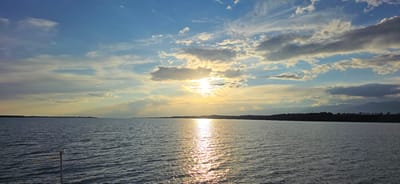 An expansive, tranquil lake stretches across the foreground, its surface rippled and reflecting the golden hues of a setting sun. The sky above is a dramatic canvas of blues and whites, with wispy clouds interspersed with larger, more dramatic formations that catch the last rays of sunlight. The horizon is marked by a dark, silhouetted landmass, likely a shoreline, with hints of trees and distant mountains adding depth to the scene. A solitary post, topped with a small metallic object and a yellow ribbon, emerges from the water on the left side of the frame, suggesting a man-made marker or structure within the natural landscape. The overall impression is one of serene natural beauty, capturing a moment of transition between day and night. The location is identified as Pristan'-Przheval'sk, Kyrgyzstan.