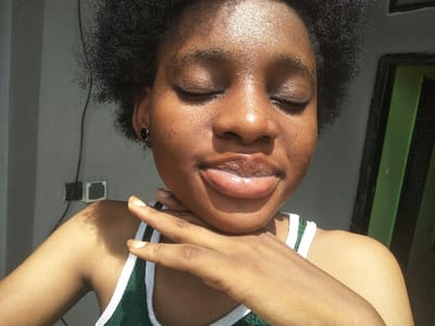 A close-up portrait depicts a young Black female with short, dark, curly hair and closed eyes, facing forward with her head slightly elevated. Her right hand supports her chin, with fingers gently curled. A small, black, circular stud earring is visible in her left ear. She wears a top featuring a green and white striped strap across her right shoulder and chest. The background to the left is a plain light gray wall, exhibiting two dark, rectangular wall-mounted fixtures and a thin black cable. To the right, a dark vertical structure with green vertical elements, consistent with a door or window frame, is partially visible. The scene is illuminated by strong, direct frontal lighting, casting distinct highlights on the subject's skin and soft shadows on the neck and shoulder. Specific geographical identification within Cotonou, Benin, is not ascertainable from the visual evidence presented in the image.