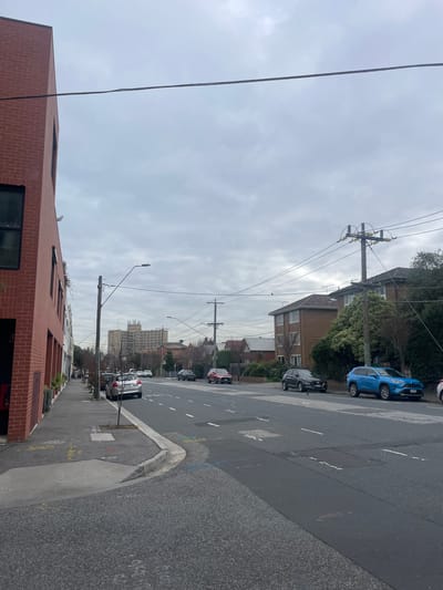 The image depicts a street scene in Balaclava, Australia. The perspective is from a low angle, looking up at a cloudy sky. A prominent power pole with multiple wires stretches across the frame, indicating an urban or suburban setting with overhead electrical infrastructure.

In the foreground, a modern brick building with large windows lines the street. Further down the street, several cars are parked along the side, including a blue SUV and a dark-colored sedan. The road is paved and marked with white lines, suggesting it is a public thoroughfare. Beyond the parked cars, more residential buildings and trees are visible, interspersed with taller structures in the distance.

The weather appears overcast, contributing to a somewhat muted and diffused light. The time of day is not explicitly clear but the lighting suggests it is daytime. No people are visible in the image, and there is no discernible activity or event taking place. The overall impression is of a quiet, everyday street scene.

There is some faint chalk or spray paint markings on the road surface near the curb, the text of which is illegible. The architectural style of the buildings, particularly the brickwork of the foreground building and the tiled roofs of others, provides some clues to the location's character.