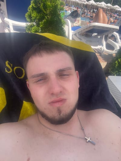 A young man with light skin, short brown hair, and a goatee lies on a sun lounger under a clear blue sky. He wears a silver cross necklace. A black towel with yellow trim is draped over him. In the background, a swimming pool is filled with people and surrounded by more sun loungers and umbrellas. Green foliage is visible to the left and right of the frame. The scene appears to be a resort or public pool area in Varvara, Bulgaria.