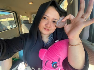 A young woman with a headscarf is taking a selfie inside a vehicle, holding a partially eaten chocolate cookie. She is wearing a denim shirt and a black headscarf that covers her hair and neck. Her left hand, adorned with rings and a bracelet, holds the cookie up to her face, while her right arm is partially visible, resting on a pink stuffed toy. The background shows the interior of the car, including car seats, windows, and sunlight streaming in, suggesting it is daytime. Outside the window, a building with a tiled roof is visible, along with trees and vegetation, indicating a suburban or rural setting. Based on the context, the location could be Kebondalem, Indonesia.