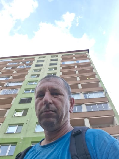 A close-up, inverted shot captures a man's face in the foreground, with his blue t-shirt and a backpack strap visible. He appears to be in his 40s or 50s, with short, dark hair and some stubble. His expression is neutral. In the background, a tall, pale green apartment building with brown accents and numerous windows dominates the scene. The building has a repeating pattern of vertical elements between sections of windows. The sky above is bright blue with scattered white clouds, suggesting it is daytime. The image has a slightly grainy quality. There is no visible text in the image. The scene appears to be an outdoor setting, possibly in a residential area. The orientation of the image suggests it was taken at an unusual angle, likely a selfie.