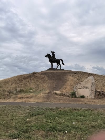 The image depicts a monumental equestrian statue silhouetted against a cloudy sky. The statue is positioned on a grassy hill, with a pathway or road running along the base of the hill. To the right of the statue, a large rock with inscribed text is visible. The text appears to be in Cyrillic script. The surrounding landscape consists of dry grass and some green vegetation in the foreground. The overall scene suggests an outdoor memorial or landmark in a somewhat arid environment. The location is identified as the city of Rogozhkin, Russia.