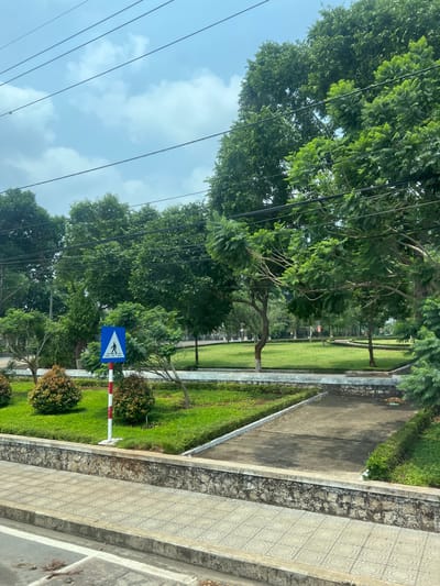 The image depicts a street scene in Phú Hòa, Vietnam. The view is taken from an elevated position. A paved walkway runs along the right side of the frame, with a patterned section closest to the viewer and a darker, plain section further away. Adjacent to the walkway is a strip of grass with small bushes and a pedestrian crossing sign. The sign is blue and white with a red and white pole. Beyond the grass strip is a paved path, separated by a low concrete barrier. A green lawn with trees stretches out into the distance. Above the trees, power lines crisscross the sky, which is partly cloudy.