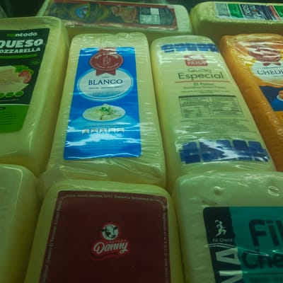 The image displays an assortment of cheese products packaged for retail sale. The primary focus is on several blocks of cheese with visible labels. On the left, a block labeled "Contodo Queso Mozzarella" is partially visible. To its right, a cheese block with a blue and white label that reads "Del Castillo Queso BLANCO SEMIDURO - SEMIGRASO - FRESCO" is prominent. Further to the right, another cheese block features a label for "Paisa Queso Especial El Paisa Semiduro Graso Fresco". A partially obscured block of orange cheese with a label indicating "CHEDDAR" is also visible on the right side. At the bottom of the image, a dark red label with the text "LACTEOS Danny" and a cow logo is affixed to a cheese product. Another cheese product on the far right has a label with the words "Fit Cheez". The cheeses appear to be arranged on shelves or a display, typical of a supermarket or grocery store environment. The lighting is somewhat diffused, suggesting indoor illumination. The exact location in the city of Los Teques, Venezuela, cannot be determined from the visual information provided in the image itself, as no specific geographical markers or textual references to Los Teques are present on the visible packaging or within the scene's composition.