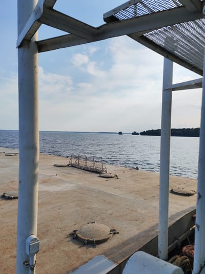 The image depicts an outdoor scene with a focus on industrial elements and a natural landscape. In the foreground, a large, weathered concrete deck is visible, marked with several circular metal hatches. Two large white metal poles with supporting beams and mesh platforms rise from the deck, framing the view. The poles and beams appear to be part of some sort of structure, possibly a ferry or industrial platform.
Beyond the deck, a calm body of water stretches to the horizon, reflecting the blue sky. The water is a deep blue and shows gentle ripples. In the distance, a line of trees forms a dark green border along the far shore, suggesting a wooded coastline. Two small boats are visible on the water, adding a sense of scale and activity to the scene. The sky is a clear blue with scattered, fluffy white clouds.
The lighting suggests it is daytime, likely late morning or afternoon, with the sun casting subtle shadows. The overall impression is one of a functional, somewhat utilitarian environment juxtaposed with the serene beauty of the natural water and sky. The scene appears to be at a dock or harbor, possibly in a rural or less developed area, given the natural surroundings. There is no visible text in the image. The activity taking place is the passive observation of a landscape from a vantage point on a structure.