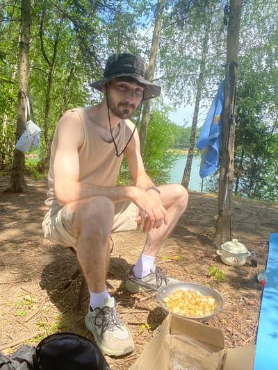 A young man wearing a camouflaged bucket hat, a light-colored tank top, and tan shorts is crouched on a folding chair outdoors. He has a beard and appears to be looking at the camera. In front of him, on the ground, is a frying pan filled with cooked food, possibly chicken. A cardboard box with plastic bags sits beside the pan. In the background, a body of water is visible, surrounded by trees. A blue towel hangs on a tree to the right of the man. The scene appears to be a campsite in or near Kaniów, Poland, given the context provided.