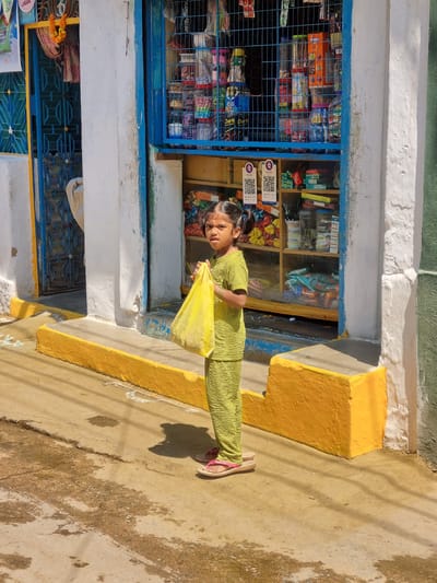 A young girl stands on a cracked concrete surface in front of a building in Pedda Kammavaripalli, India. She is positioned slightly right of center, facing the camera directly, wearing a textured, light green two-piece outfit and pink flip-flops. Her dark hair is styled in two pigtails, and a red mark is visible on her forehead. She holds a yellow plastic bag with both hands in front of her midsection.
Behind the girl, the building features a white painted facade with blue accents around openings. To her right, a shop window with blue metal grating displays an assortment of packaged goods, including food items and household products, on interior shelves. Two PhonePe QR code payment signs are clearly visible within the shop display. To her left, a blue metal gate with a geometric pattern is partially open, revealing a dark interior space. A marigold garland hangs above this gate. A yellow painted curb or raised platform runs along the base of the building, separating the storefront from the ground. Faint white chalk-like patterns are visible on the concrete near the curb. The ground shows patches of discoloration and distinct shadows from overhead objects and the girl herself, indicating bright sun exposure.