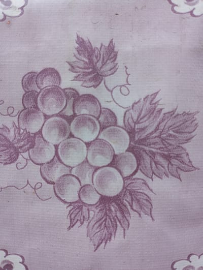 The image displays a close-up of a fabric with a printed pattern of grapes and leaves in a shade of purple. The fabric has a textured appearance, and the print is slightly faded, suggesting it might be old or worn. There are also small, scattered dark specks on the fabric, possibly dust or imperfections. The pattern is repeated, with portions of other clusters of grapes and leaves visible at the edges of the frame.
This image does not depict any scene in the city of Voznesenske, Ukraine, nor does it contain any people, actions, or interactions. It is a static image of a textile print.