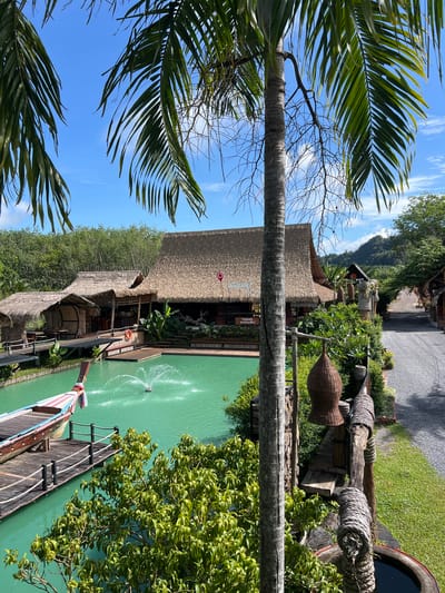 This image depicts a tranquil tropical scene in Tambon Pa Kae Bo Hin, Thailand. The setting features traditional thatched-roof buildings nestled amongst lush green vegetation and a vibrant, turquoise body of water. In the foreground, large palm fronds frame the view, suggesting a sunny day with a clear blue sky dotted with wispy clouds.
The water is calm, with a fountain creating a gentle spray, and a single person is visible in the distance, seemingly enjoying the serene environment. A long, wooden boat with colorful decorations is moored to a wooden dock, adding a touch of cultural authenticity. The architecture of the buildings, with their natural materials and open designs, emphasizes a connection with the surrounding environment. The overall atmosphere is one of peaceful relaxation and natural beauty.
There are no prominent signs with visible text, but a small red marker pin is visible on one of the buildings, potentially indicating a point of interest or location. The time of day appears to be daytime, with bright sunlight casting distinct shadows. The weather seems pleasant and warm, typical of a tropical climate. The location cues, such as the vegetation, architecture, and the boat style, strongly suggest a Southeast Asian tropical destination.