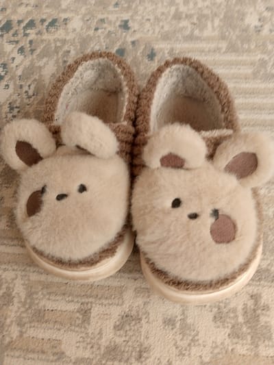The image shows a pair of fluffy, beige, bear-shaped slippers resting on a textured rug. The slippers have round ears with brown accents and embroidered facial features consisting of small black dots for eyes and a dash for a mouth. One slipper also has a brown accent on its cheek. The rug appears to be a light brown or beige color with a subtle pattern of darker lines and shapes. The slippers are positioned side-by-side, with the left slipper slightly overlapping the right. The overall impression is one of comfort and cuteness. There is no indication of the city of Prigorognoye, Kyrgyzstan in the image.
