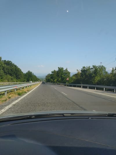 The image shows a view from inside a vehicle traveling on a highway in Yagoda, Bulgaria. The highway is paved and has multiple lanes, with a guardrail separating the oncoming traffic from the shoulder. Lush green trees line both sides of the road, indicating a rural or semi-rural setting. The sky is clear and blue, with a faint appearance of the moon or a distant celestial body. Several vehicles are visible on the highway ahead, suggesting moderate traffic. The dashboard of the vehicle is visible in the foreground, framing the road ahead.