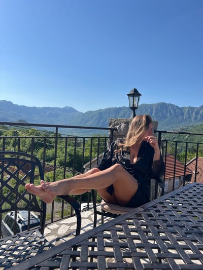A woman in a black robe sits on a black wrought iron chair on a balcony overlooking a mountainous landscape under a clear blue sky. A black wrought iron table sits beside her. A street lamp stands next to the balcony. The background includes buildings with red tile roofs, trees, and mountains. The location is likely Sotonići, Montenegro.