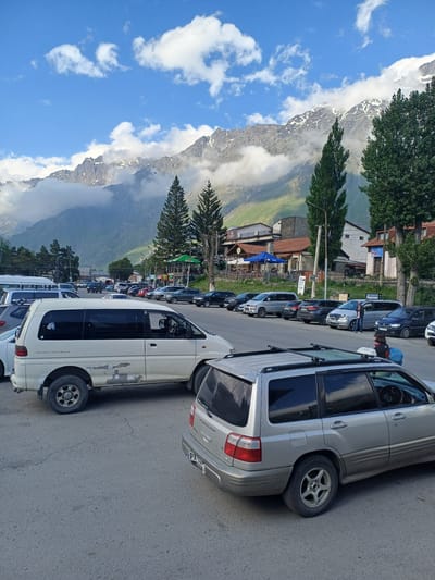 A parking lot in Stepantsminda, Georgia is filled with a variety of vehicles, including vans and SUVs. In the background, there are several buildings that appear to be shops or restaurants, with outdoor seating areas visible. Beyond the buildings, tall evergreen trees are scattered, and in the distance, the majestic snow-capped peaks of the Caucasus Mountains rise up, partially obscured by soft, white clouds. The sky is a clear blue, with a few scattered white clouds. The ground is paved asphalt, showing some cracks and wear. A few people are present, some standing near the vehicles and others walking. One person, wearing a blue backpack, is standing next to a silver SUV. The lighting suggests it is daytime, with the sun casting shadows. The overall scene depicts a busy parking area in a scenic mountain town.
