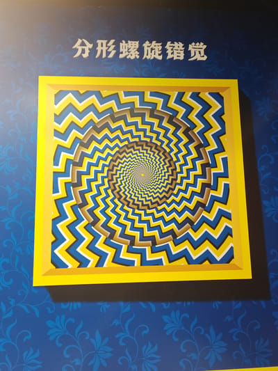 This media file captures a framed optical illusion artwork on a blue patterned wall. The artwork itself is a circular, spiraling pattern of zig-zagging lines in yellow, blue, and grey, creating a sense of depth and movement. The illusion is further enhanced by a bright yellow frame surrounding the artwork.
Above the artwork, white Chinese characters are visible on the blue wall, reading "分形螺旋错觉". This translates to "Fractal Spiral Illusion."
The background wall is a vibrant blue, decorated with a subtle, repeating floral or vine-like pattern in a slightly darker shade of blue. The lighting suggests an indoor setting, possibly a museum or exhibition space, given the presence of the artwork and the way it's displayed. There are no people or specific environmental cues like weather or time of day visible in the frame. The focus is entirely on the art and its presentation. The overall impression is one of visual intrigue and a deliberate attempt to create a captivating effect through art and design.