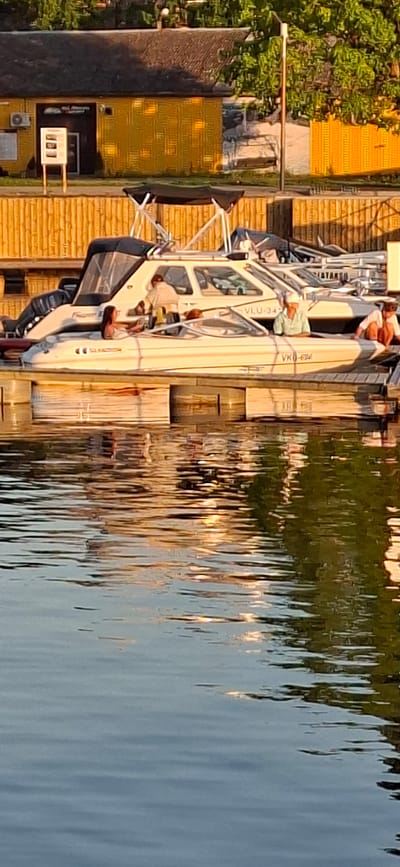 The image shows a waterfront scene in Pärnu linn, Estonia. Several boats are docked at a wooden pier. In the foreground, the rippling surface of the water reflects the sunlight and the surrounding environment. On the boats, there are at least three people visible. One person, wearing a light-colored shirt, is leaning over the edge of a boat, appearing to interact with someone inside. Another person, wearing a green and white striped shirt and a white cap, is looking forward from the cockpit of a boat. A third person, wearing an orange shirt, is seated on the edge of a boat. In the background, there is a yellow building with a dark roof and some trees. The building appears to have an air conditioning unit on its side and a sign with information posted outside. The overall atmosphere is relaxed, suggesting a recreational setting.