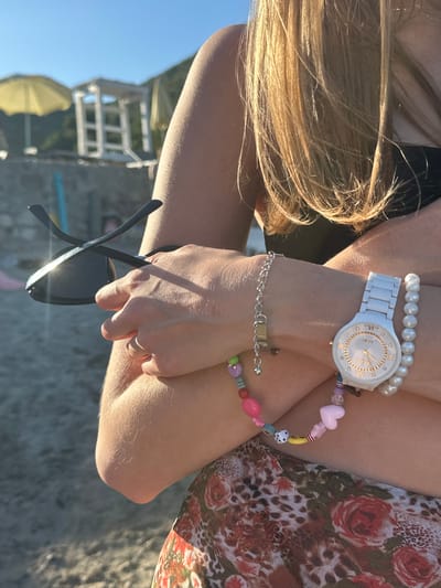The image depicts a close-up, slightly angled view of a person on a sunny beach in Lastva Grbaljska, Montenegro. The upper right portion of the image shows the person's blonde hair, illuminated by sunlight, and a section of a black top. The person's arms are crossed, with the right hand holding a pair of black sunglasses that exhibit a strong sun glare on the lens. The left arm is adorned with multiple accessories: a silver-colored chain bracelet with a small charm, a white Rado wristwatch with gold-toned Roman numeral markers (XII, VI) and small diamonte accents on the face, a white pearl bracelet, and a colorful beaded bracelet. This beaded bracelet features various shapes including pink hearts, a white dice, a yellow smiley face, and other multicolored spherical and cylindrical beads. The skin on the arms and hands shows fine hair, highlighted by the sunlight. Below the arms, the person wears clothing with a distinct pattern combining red roses and a leopard print on a mesh-like fabric. The background, largely out of focus, reveals a bright blue sky, blurred yellow beach umbrellas, and a white, multi-tiered structure resembling a lifeguard stand. The ground beneath is sandy or pebbled beach material.