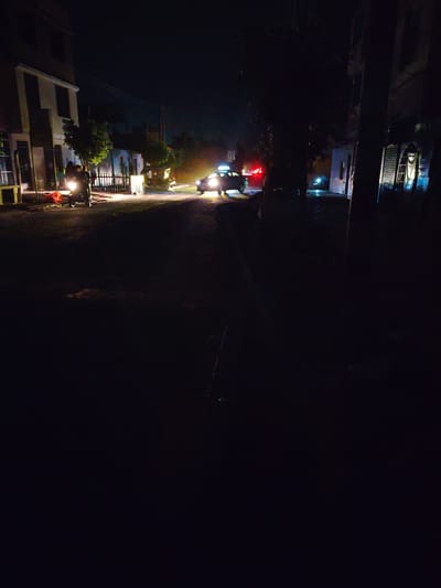 A dark street scene at night, likely in a residential area of Trujillo, Peru. A motorcycle with two people on it is parked on the left side of the street. A car with its headlights on is driving down the street. The environment includes buildings on both sides of the street. Illumination is minimal, with light sources primarily emanating from vehicle headlights and indistinct street lighting. The scene's overall visibility is poor due to the darkness.