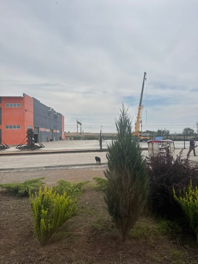 The image depicts an outdoor industrial or construction site under an overcast sky. In the foreground, there are several meticulously arranged bushes and plants, including a tall, slender evergreen tree in the center. To the left, a building with an orange facade and dark gray walls is visible, featuring a row of windows on the orange section. In front of the building, two large, heavy industrial equipment pieces, possibly part of a crane or loading mechanism, are positioned on railway tracks.
Further into the scene, a large yellow crane is actively at work, its arm extended upwards. Near the crane, a small, translucent shelter with a table and chairs inside is present. A person in camouflage pants and a dark shirt is walking away from the shelter towards the right edge of the frame. A small black dog is visible in the mid-ground, near the industrial equipment. In the background, a fence line with barbed wire runs across, and beyond it, a landscape of rolling hills covered in vegetation is observed. The overall environment suggests an active industrial area, likely in the city of Jıbek joly, Kazakhstan, given the context.