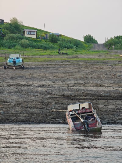 The image depicts a waterfront scene in the village of Potapovo, Russia. In the foreground, a small, red and white boat with an outboard motor is partially submerged in the water and leaning against the muddy shore. The boat appears to be in disrepair, with peeling paint and visible wear and tear. Its propeller is submerged. In the middle ground, a light blue boat with a motor is resting on a trailer on the dry, muddy bank. The bank is a mixture of dark soil and small pebbles. In the background, a grassy slope is dotted with green bushes and small trees. Several small buildings, including a white structure with a dark roof and windows, are visible on the hillside. A dirt path or ramp leads up the slope. Two figures are visible on the slope, one walking up the ramp and another standing further back. The sky is overcast and pale. The overall impression is one of a rural, functional settlement by a body of water, with evidence of a somewhat harsh environment.