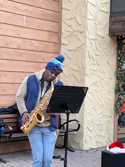 A man plays the saxophone while seated on a bench. He wears a blue beanie, sunglasses perched on the hat, and a blue and beige jacket. A black music stand is positioned in front of him. The background features a textured wall, a window decorated with greenery, and a glimpse of a Santa hat. The setting appears to be outdoors, possibly a public space or venue.