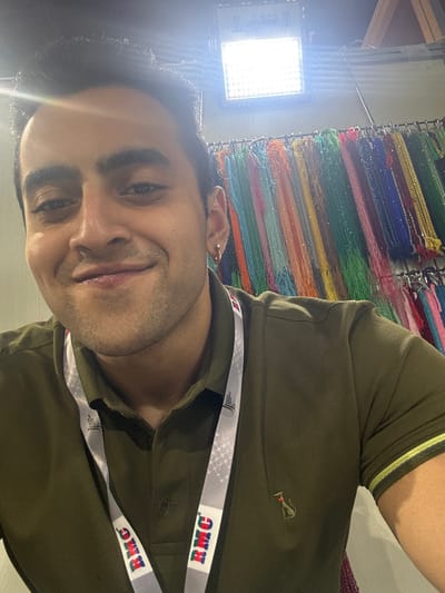 A close-up photograph features one adult male subject, estimated to be in his 20s or 30s, captured in a frontal portrait. The subject has dark hair, dark eyes, and light facial stubble, and is smiling directly at the camera. He is wearing a dark olive green short-sleeved polo shirt, which includes a small, light-colored embroidered emblem on the left chest. A white lanyard with a grey and white checkered pattern, displaying "RMC" logos in red and blue text, is worn around his neck. A small, clear stud earring is visible in his left earlobe. The background consists of numerous vertically suspended strands of colorful beads or small decorative elements, arranged in various hues such as green, blue, yellow, orange, and red, suggesting a retail environment like a market stall or exhibition booth for jewelry or craft items. A bright, square artificial light source is visible in the upper right background, creating a lens flare. The scene is situated within the city of Jaipur Municipal Corporation, India.