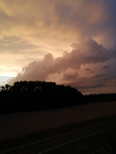 The image captures a dramatic twilight sky over a rural landscape. The sky is filled with voluminous clouds, illuminated by the setting sun, casting a warm, golden-orange hue across the scene. The clouds range from soft, ethereal wisps to darker, more ominous formations. Silhouetted against this vibrant sky is a dense line of trees forming a dark, horizontal band across the middle of the frame. Below the treeline, a dark field stretches towards a road with white markings, indicating it is likely a public road. In the far distance, beyond the field and trees, faint structures of buildings or infrastructure can be discerned, suggesting the proximity of a town or settlement. The overall impression is one of natural beauty and a transition from day to night. The image is not specific to Ecublens, Switzerland, as there are no identifiable landmarks or features that place it in that specific location.