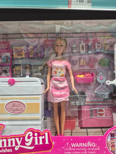 The image shows a toy playset, likely a Barbie doll and accessories, packaged in a clear plastic box. The playset appears to be a grocery store or market theme, with miniature grocery items, a cash register, a shopping cart, and a doll dressed as a cashier or shopper. The background suggests a retail environment, with shelves and products visible behind the main playset. The doll is wearing an apron with a cat graphic and the word "welcome" printed on it. The packaging features the text "Funny Girl" and a "WARNING: CHOKING HAZARD—Small parts. Not for children under 3 years." The location in the city of Proshyan, Armenia, is not evident from the image itself; this information is external to the visual content.