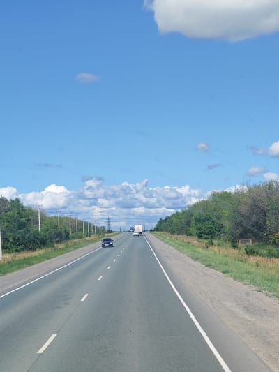 The image captures a view from a vehicle traveling on a paved highway under a clear blue sky with scattered white clouds. The road is a two-lane highway, with a white solid line on the left and dashed white lines dividing the lanes. The highway is flanked by green trees and vegetation on both sides. There are utility poles visible along the left side of the road. In the distance, several vehicles, including cars and trucks, are visible on the highway. The scene suggests a rural or semi-rural setting, possibly in Maloe Tomylovo, Russia, given the context.