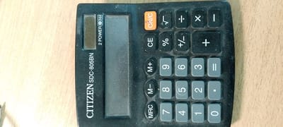 The image shows a black Citizen SDC-805BN calculator placed on a light-colored wooden surface. The calculator is oriented vertically and is slightly dusty. The display screen is dark. Various buttons are visible, including numerical keys, arithmetic operators, and function keys such as "ON/C", "CE", "%", "+/-", "M+", "M-", "MRC", and "=". The image is a close-up of the calculator and the wooden surface, with no other objects or people present. The exact location of Zarechny, Russia, is not discernible from the visual information provided in the image.