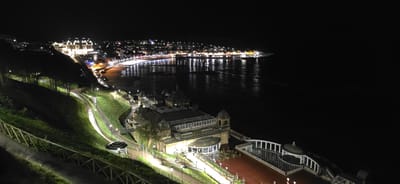At night, an elevated long shot focuses on the architecture and landscape of Scarborough, United Kingdom. The cityscape is partially visible in the top left, illuminated by streetlights and building lights. The lights reflect on a body of water separating the city from a dark open space.
Prominently displayed below and to the right of the cityscape is an ornate building with a glass roof and multiple dome-shaped structures. Pathways lit by lampposts lead to the building and other smaller structures. The bottom left shows a hillside covered in greenery with a wooden fence lining a path.
The environment is dark overall. The focus is the contrast between artificial lighting and natural darkness. No people are visible.