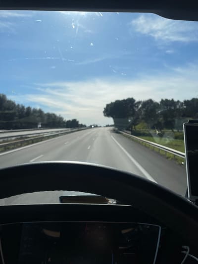 The image depicts a driver's perspective from within a vehicle traveling northbound on a multi-lane highway near Tavel, France, under a clear blue sky with scattered white clouds and a high sun causing lens flare. The asphalt road features white lane markings and stretches straight into the distance, reflecting sunlight. Guardrails line both sides of the highway. To the left, a line of trees and foliage is visible beyond the guardrail. To the right, green vegetation transitions to a cluster of dark green trees, including one distinctively shaped tree. The foreground shows the upper arc of a dark steering wheel. Below the steering wheel, a digital instrument panel displays unreadable icons and segments. An object, possibly a smartphone, rests flat on the dashboard between the steering wheel and the instrument panel. On the far right, a vertically mounted electronic device is partially visible with a white cable extending downwards. The windshield exhibits minor dust and streak marks. No individuals are visible.
