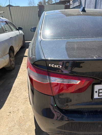 The image shows the rear of a black Kia Rio parked in an outdoor environment. The background indicates a likely urban or semi-urban setting in Зуй, Russia, with buildings and another vehicle partially visible. The Kia Rio is dusty, and the focus is on its rear taillight and the 'RIO' badge. The ground is concrete or similar material.