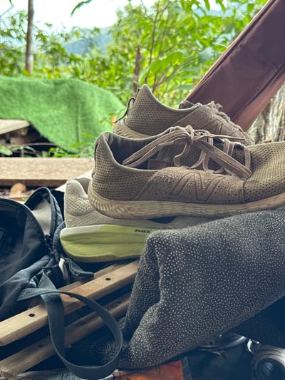 The image depicts a collection of items, including a pair of tan-colored running shoes with laces tied, hanging among other objects. To the right of the shoes, there's a gray fabric with a pattern of white dots. Further to the right is what appears to be a wooden folding chair and a dark-colored backpack.

In the background, visible through the gaps between the hanging items, there is greenery suggesting an outdoor environment. The presence of a brown awning-like structure above hints at a sheltered or covered area.

Given the location as Moo 7, Thailand, without additional context, it is difficult to provide further forensic information.