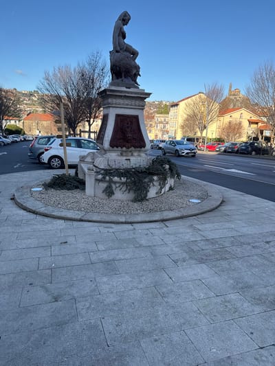 The image displays the "L'Enfant au Bélier" (Child with Ram) fountain situated in the Place du Breuil, Le Puy-en-Velay, France. The central subject is a weathered stone fountain featuring a partially draped female nude statue seated atop a large ram or similar horned animal. The statue rests on an ornate, multi-tiered pedestal adorned with carved bas-relief panels, one of which depicts a figure. Evergreen boughs are draped around the lower basin of the fountain, which is encircled by light-colored gravel contained within a stone curb. The fountain stands on a paved pedestrian area composed of light grey rectangular stone tiles.
To the right, a multi-lane asphalt road shows a silver Renault Clio hatchback in motion, driving away from the viewer. Several parked vehicles, including red and dark-colored cars, line the far side of the road. To the left, a paved parking area contains multiple parked cars, including silver and white hatchbacks. A wooden post structure is partially visible near the parked cars on the left.
The background reveals the urban landscape of Le Puy-en-Velay, characterized by multi-story buildings with various facades and numerous bare deciduous trees. Prominently in the distant upper right, the reddish-brown statue of Notre-Dame de France atop the Rocher Corneille, a volcanic plug, is visible. The sky is clear and bright blue, indicating daylight conditions with the sun casting long shadows, consistent with winter or early spring. No individuals are present in the scene.