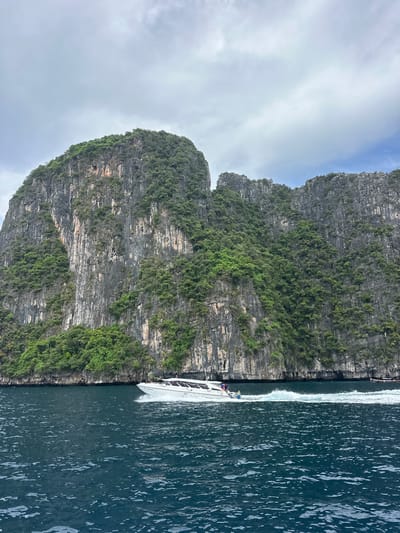 A high-angle, full shot captures a speedboat moving to the left on dark blue water. The boat is white with a dark stripe and has several passengers visible. Behind the boat, a steep, rocky cliff face covered in lush green vegetation rises dramatically from the water. The cliff has vertical striations and appears weathered. The sky above is overcast with gray clouds. The scene is located at Haad Hin Khom, Thailand.